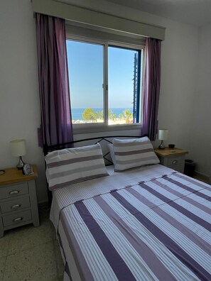 A bedroom with a bed, two bedside tables, a window with curtains, and a view of the sea.