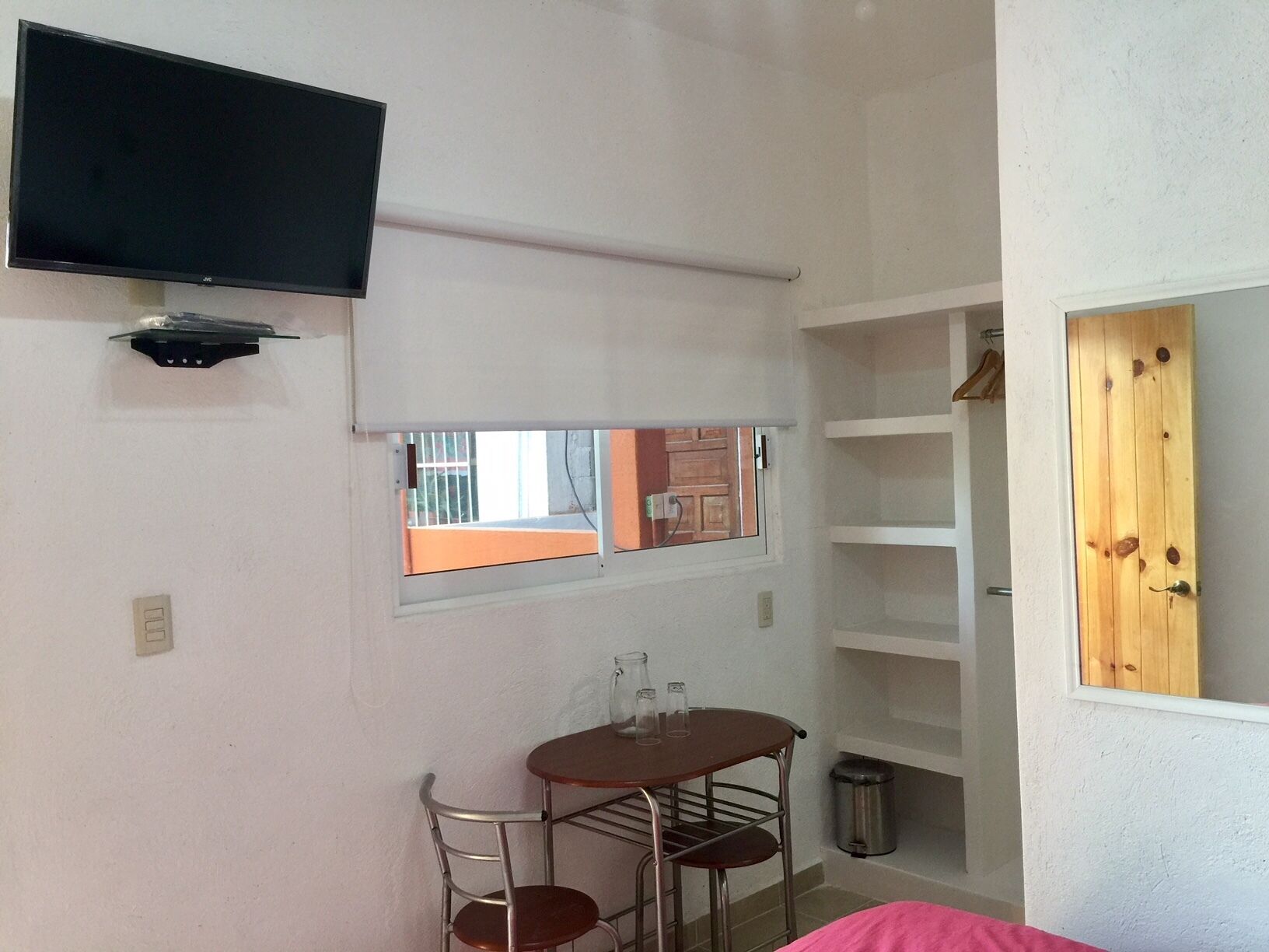 Una habitación pequeña con un televisor montado en la pared, una ventana con vistas al exterior, una mesita con sillas y una puerta de madera.