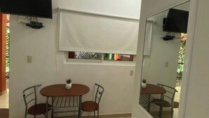A small, well-lit room with a bed, a dining table with chairs, a window with blinds, and a flat-screen TV mounted on the wall.