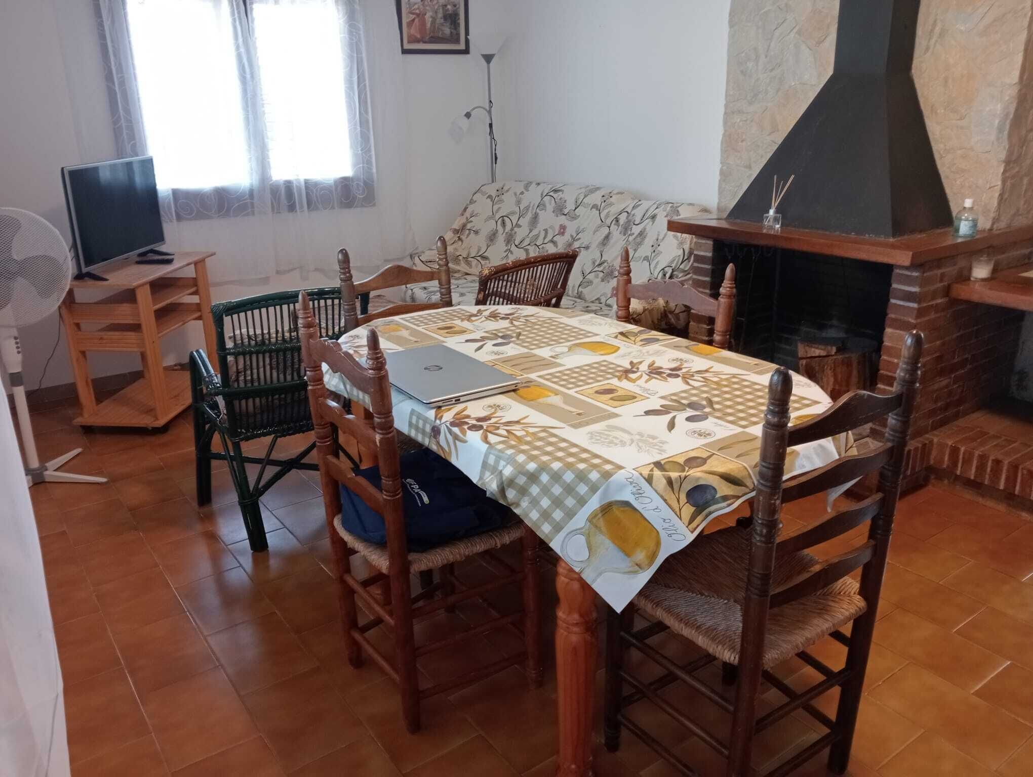 A dining area with a table set for four, surrounded by wooden chairs, a laptop on the table, and a fireplace in the background.