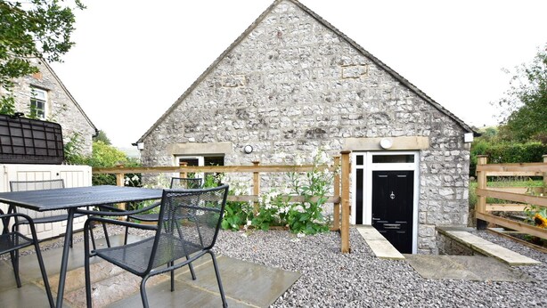 A stone building with a black door, a patio with a table and chairs, and a wooden fence. - Recently renovated 2 bedroom annexe with open plan living room and kitchen. (Stoney Middleton)