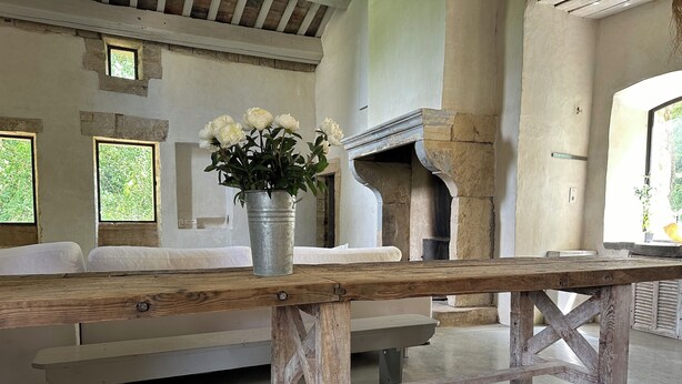 A rustic wooden table with a metal bucket of white flowers, a stone fireplace, and a bench with white cushions in a room with large windows. - In Uzès, quiet, loft in independent 18th century mill with 15mx3m swimming pool (Uzès)