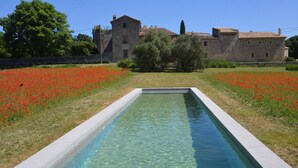 A rectangular pool with clear water, surrounded by a grassy area with red flowers, and a stone building in the background. - In Uzès, quiet, loft in independent 18th century mill with 15mx3m swimming pool (Uzès)