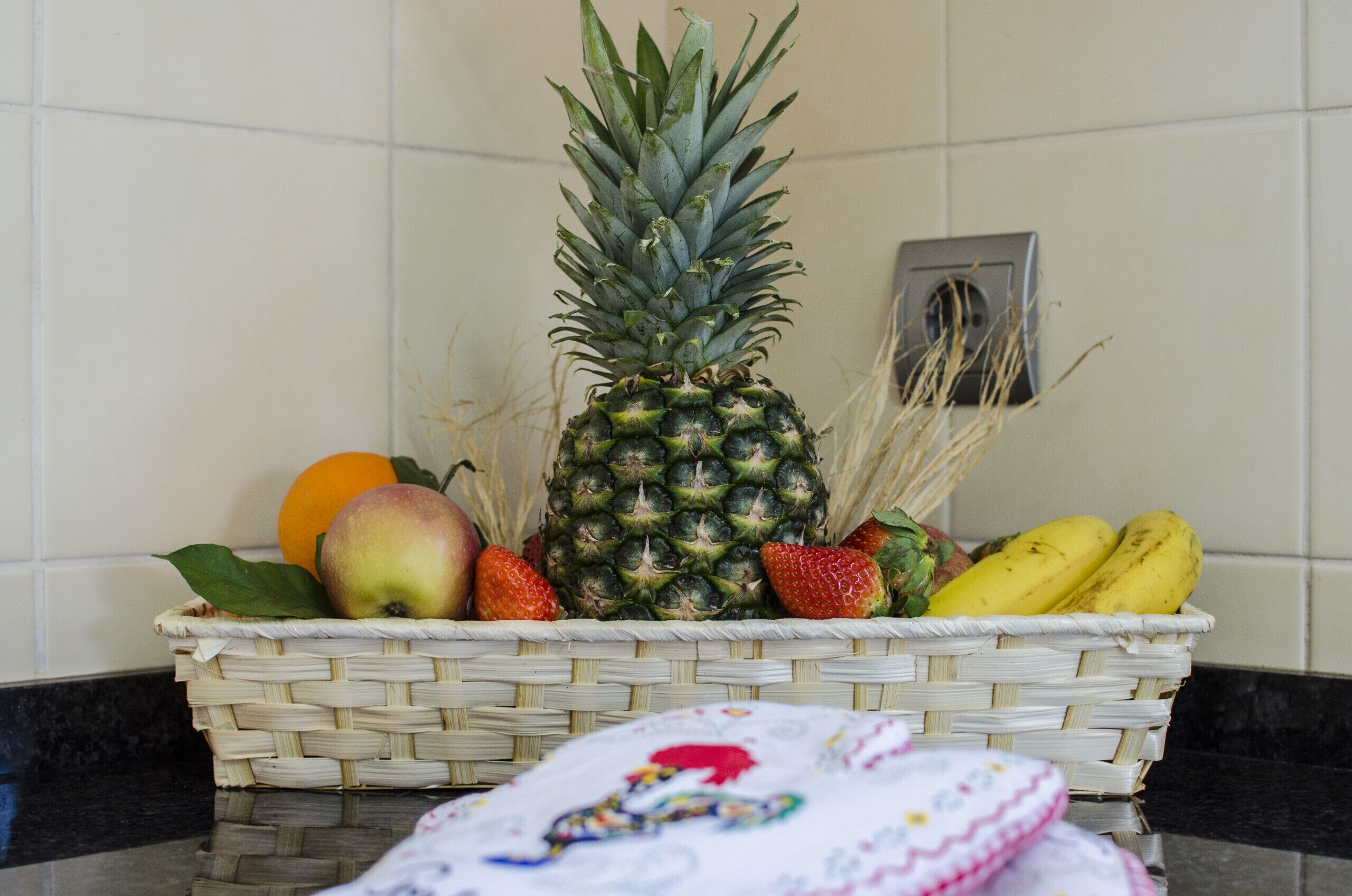 Una canasta de mimbre con una piña, fresas, plátanos y una manzana sobre una encimera de cocina.