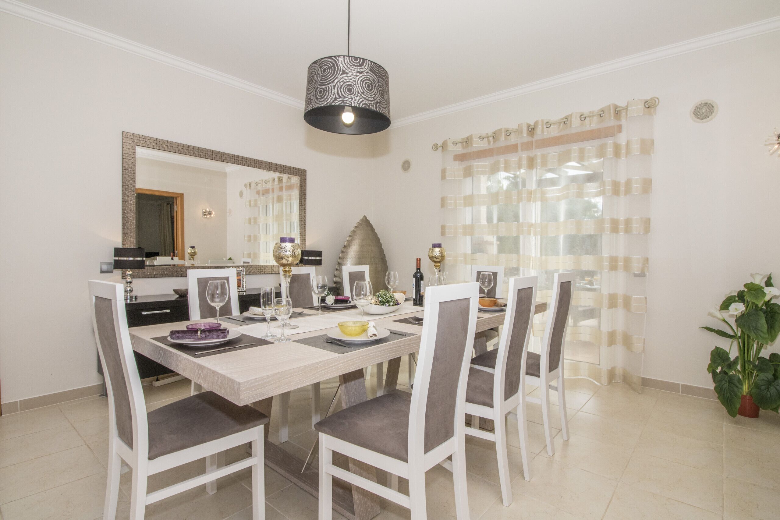 Uma sala de jantar com uma mesa comprida, cadeiras, um candelabro, um espelho e um vaso com planta.