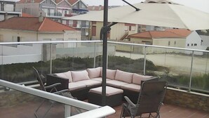 Un balcón con barandilla de vidrio, un paraguas color beige, una silla negra y un sofá rosa.