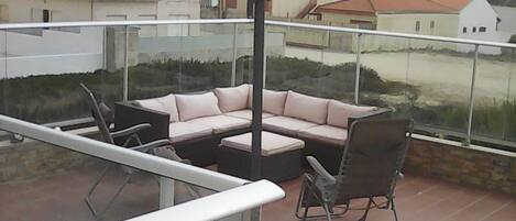 Ein Balkon mit Glasgeländer, einem beige farbenen Sonnenschirm, einem schwarzen Stuhl und einem rosafarbenen Sofa.