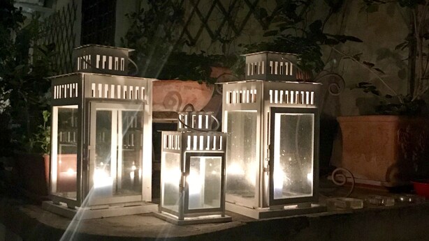 A row of four white lantern-style lights on a ledge, with a lattice fence and potted plants in the background. - Count Checco. A heaven in the rooftops of Rome few steps away from the pantheon (Roma)