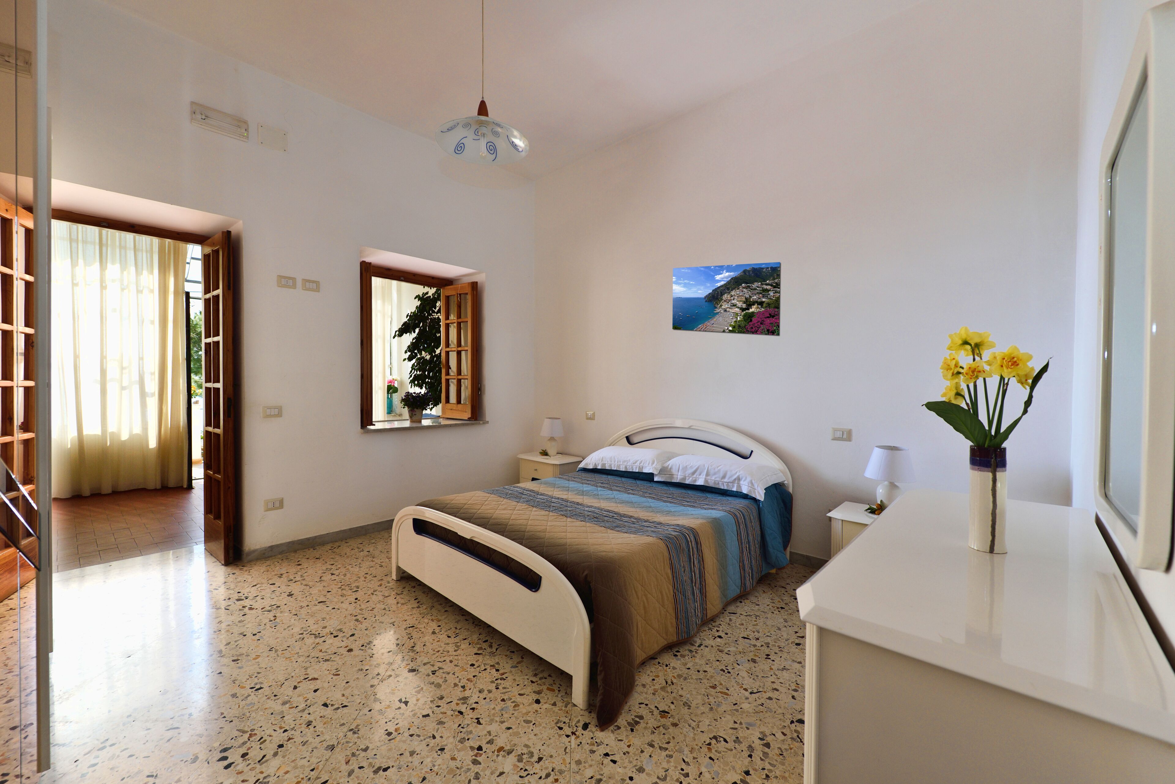 Un dormitorio con cama, mesitas de noche, un ventanal con vistas, una puerta y un cuadro colgado en la pared.