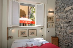 A bedroom with a bed, a window with an open shutter, a red umbrella, and a stone wall.