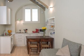 Una cocina compacta con armarios blancos, una mesa de comedor de madera y una sola silla. Una pequeña ventana con persianas blancas se encuentra sobre el fregadero.
