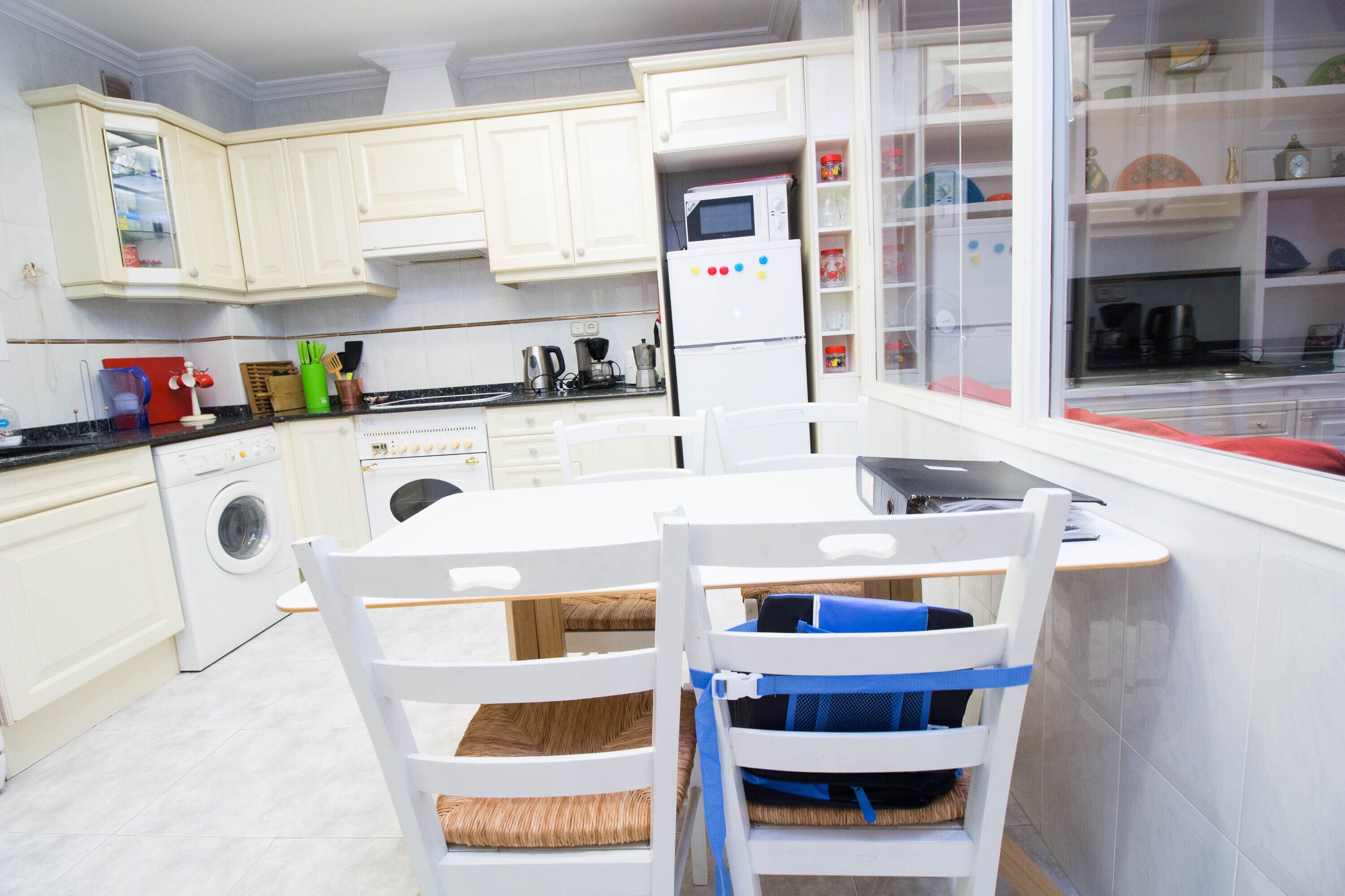 Une cuisine moderne avec des meubles blancs, un four encastré et un coin repas avec des chaises blanches.