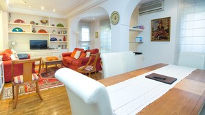 Un salon avec une table à manger en bois, des canapés rouges et blancs, un téléviseur et une bibliothèque ornée d’assiettes décoratives.
