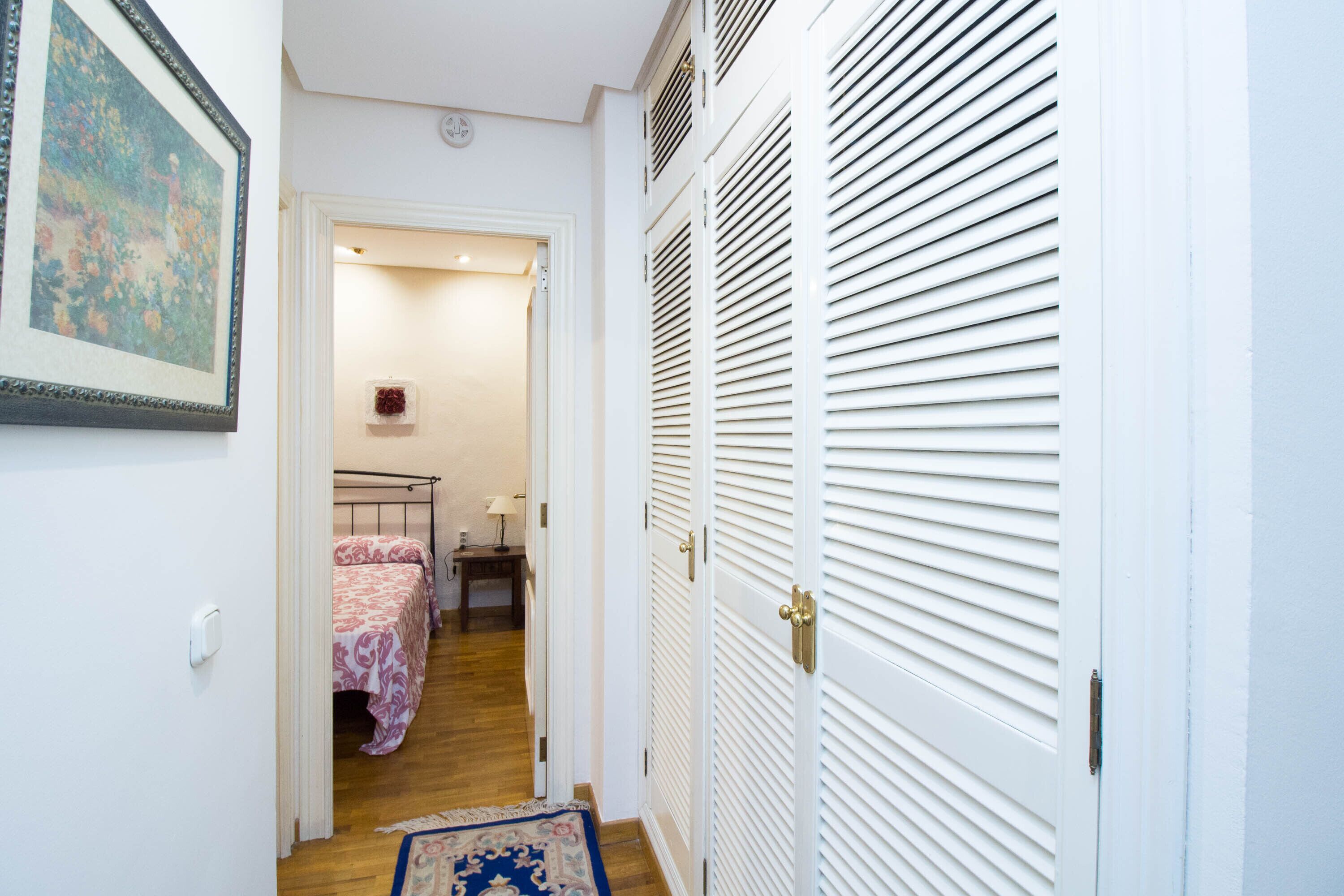 A narrow hallway leading to a bedroom with a bed, bedside table, and a small rug.
