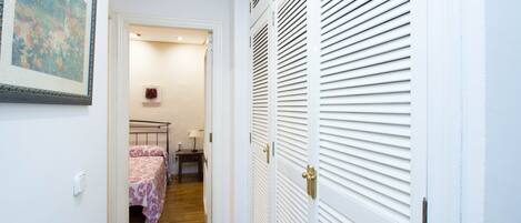A narrow hallway leading to a bedroom with a bed, bedside table, and a small rug.