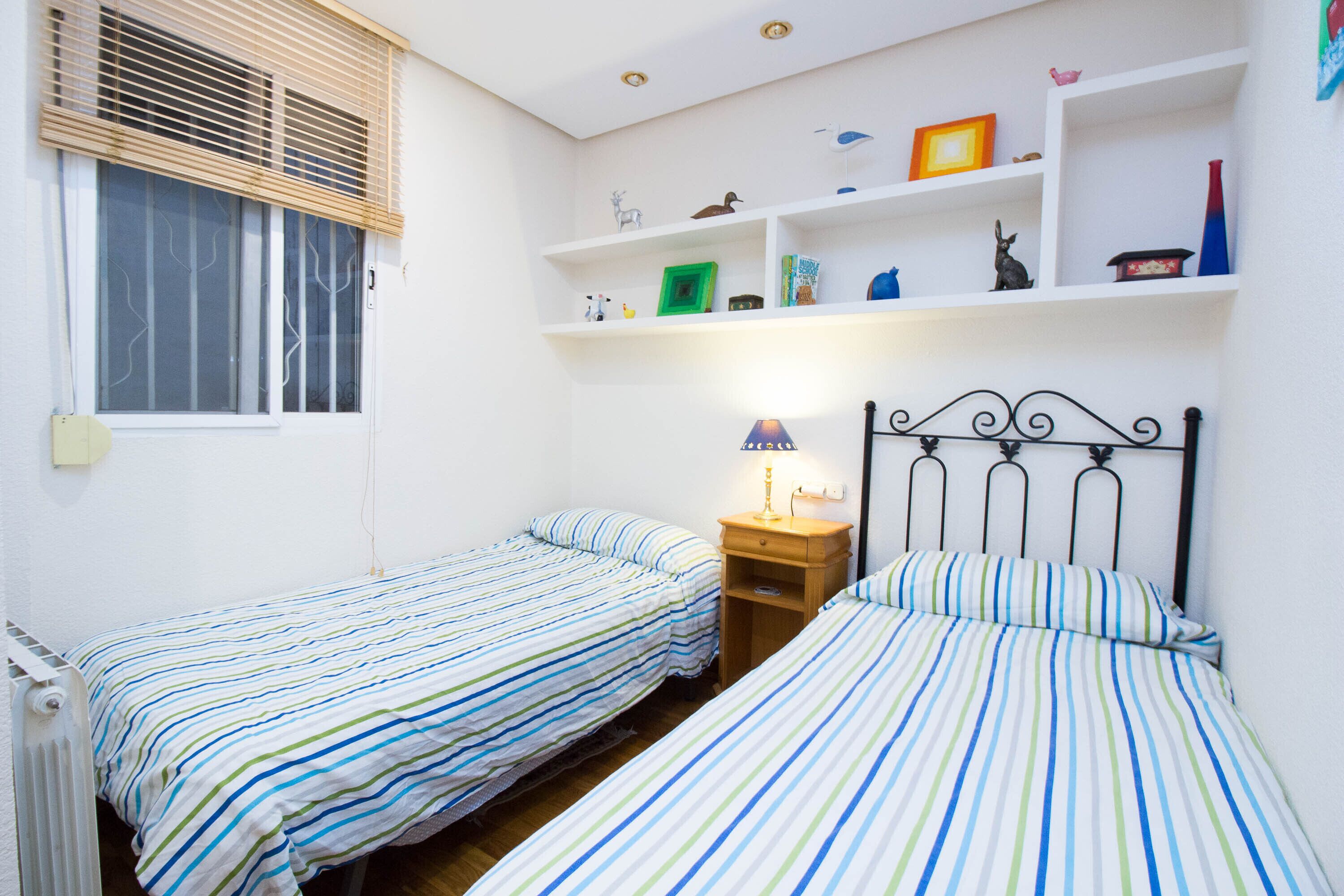 A small, clean room with two single beds, a nightstand with a lamp, and a shelf with various items.