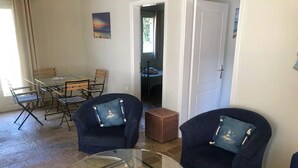 Ein Wohnzimmer mit zwei Sesseln, einem Glastisch, einem Esstisch mit Stühlen und einem Korbstuhl.