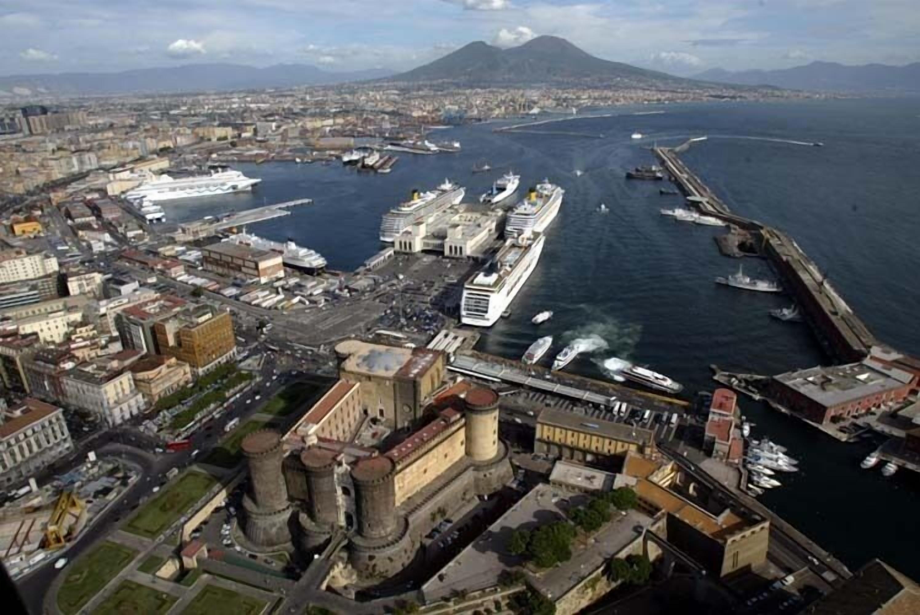 Un paisaje costero urbano con un puerto destacado donde están amarrados grandes cruceros, una fortaleza histórica y montañas al fondo.