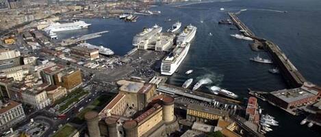 Un paisaje costero urbano con un puerto destacado donde están amarrados grandes cruceros, una fortaleza histórica y montañas al fondo.