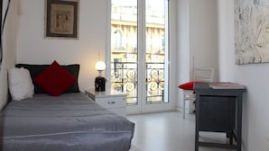 A bedroom with a bed, a nightstand, a lamp, a window with a view of buildings, and a door. - Naples city of the Sun and the Sea and 2500 years of history awaits you (Napoli)