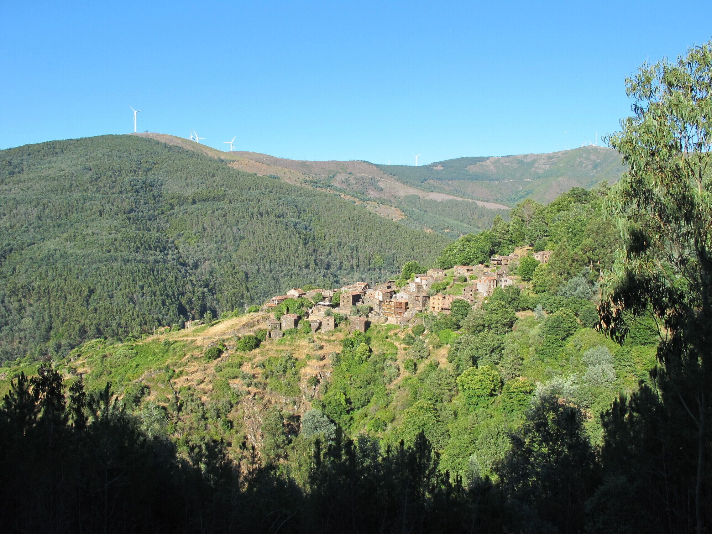 A hillside village with traditional stone buildings, surrounded by dense forest and hills.