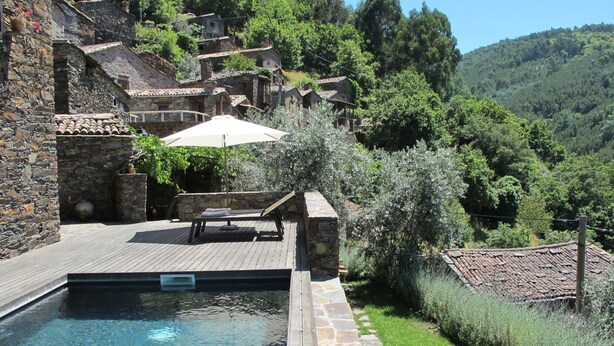 Ein Pool mit Steinmauern, eine Lounge-Bereich mit Tisch und Stühlen sowie Blick auf ein Hügeldorf.