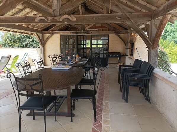 An outdoor patio with a wooden table and chairs, a grill, and a view of greenery.