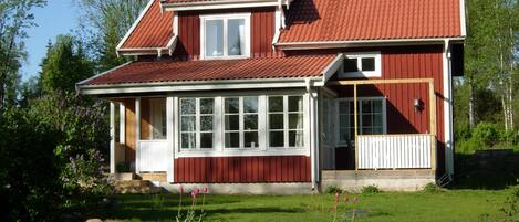 Een tweelaags huis met een rood dak en witte omlijsting, met grote ramen en een stenen fundering.
