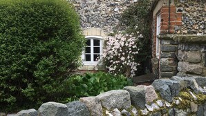 Un mur de pierre avec une petite fenêtre et un arbuste aux fleurs roses.