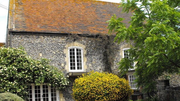 Un bâtiment en pierre avec un toit en tuiles, une fenêtre et un jardin bien entretenu, agrémenté de fleurs jaunes et d’une haie.