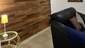 A living room with a wooden panel wall, a black leather sofa with cushions, a wicker chair, and a small table with a lamp. - Flic En Flac: Furnished 2 bedrooms  1 min walk from beach with WIFI & Nespresso (Flic- en-Flac)