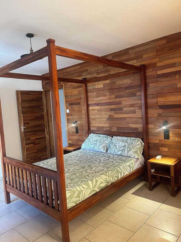 A four-poster bed with a leaf-patterned bedspread, a nightstand with a lamp, and a wooden wall paneling. - Flic En Flac: Furnished 2 bedrooms 1 min walk from beach with WIFI & Nespresso (Flic- en-Flac)