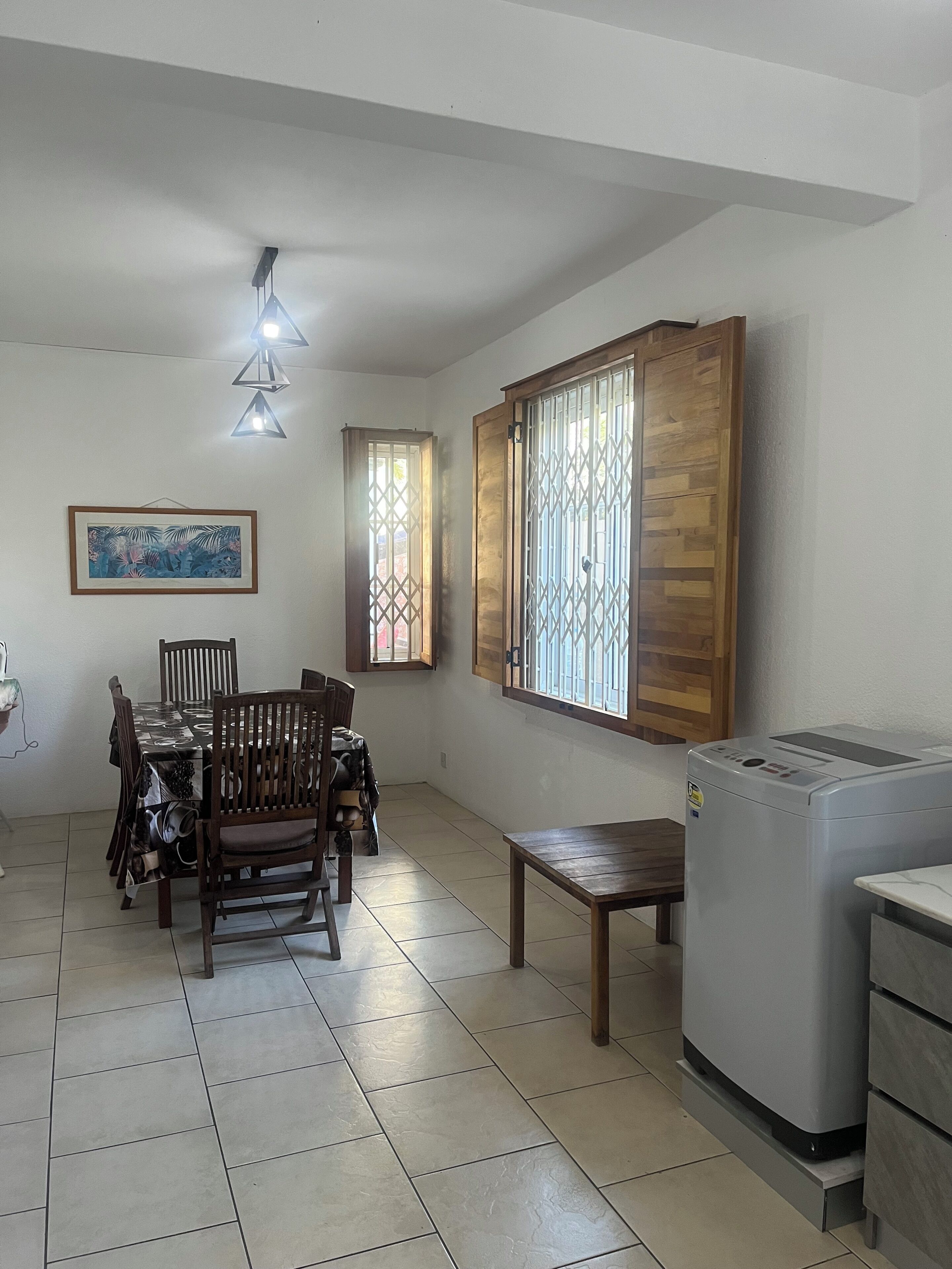 Un coin repas avec une table et des chaises, une fenêtre avec des barreaux, un luminaire au plafond et un réfrigérateur.