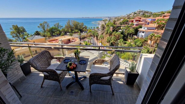 Un balcon agrémenté de fauteuils et d’une table en osier, offrant une vue sur une ville côtière avec ses bâtiments et ses palmiers.