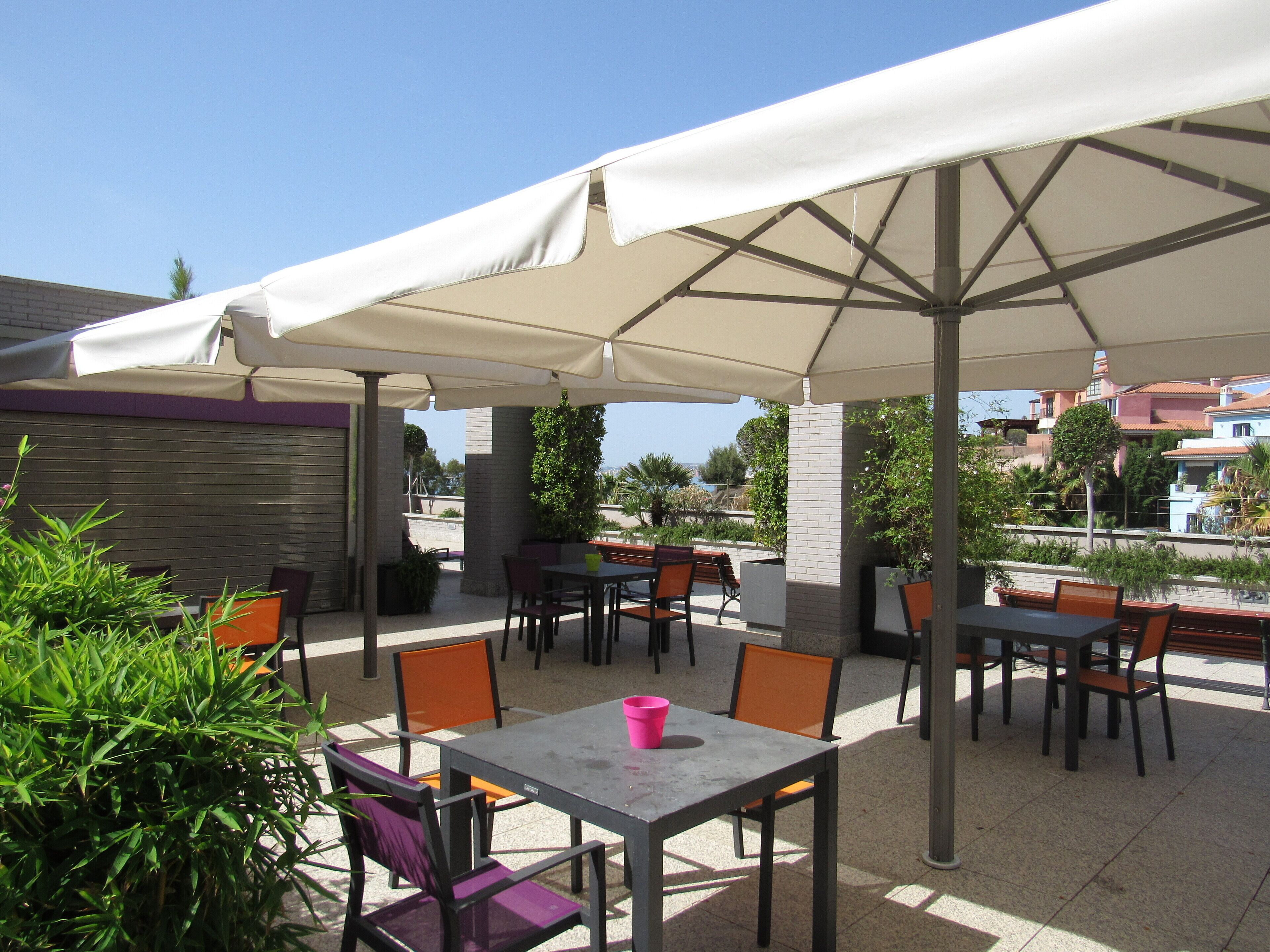 Un espace extérieur aménagé en terrasse, avec des tables et des chaises placées sous de grands parasols, entouré de plantes en pot et offrant une vue sur les bâtiments en arrière-plan.