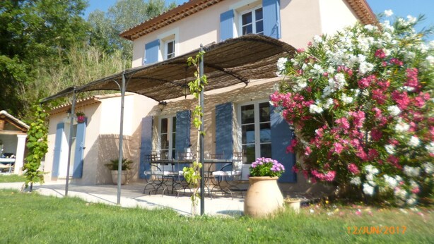 Ein zweistöckiges Haus mit einer überdachten Terrasse, einem Essbereich und einem Garten mit rosa und weißen Blumen.