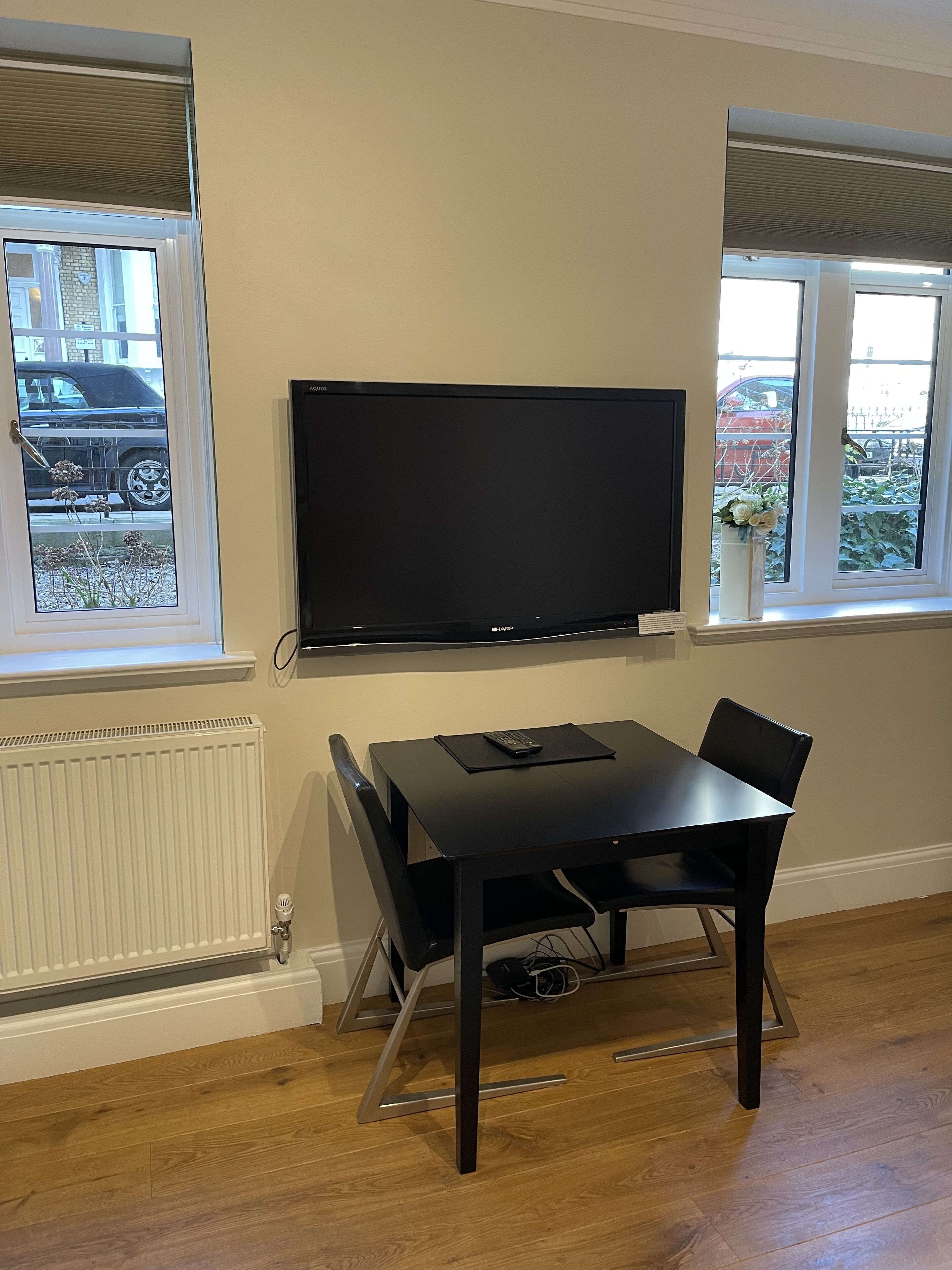 A room with a flat-screen TV mounted on the wall, a black dining table with chairs, and two windows with views of parked cars and greenery outside.