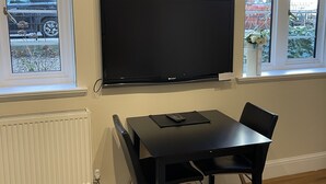 Una stanza con TV a schermo piatto fissata al muro, un tavolo da pranzo nero con sedie e due finestre che danno su auto parcheggiate e vegetazione all'esterno.