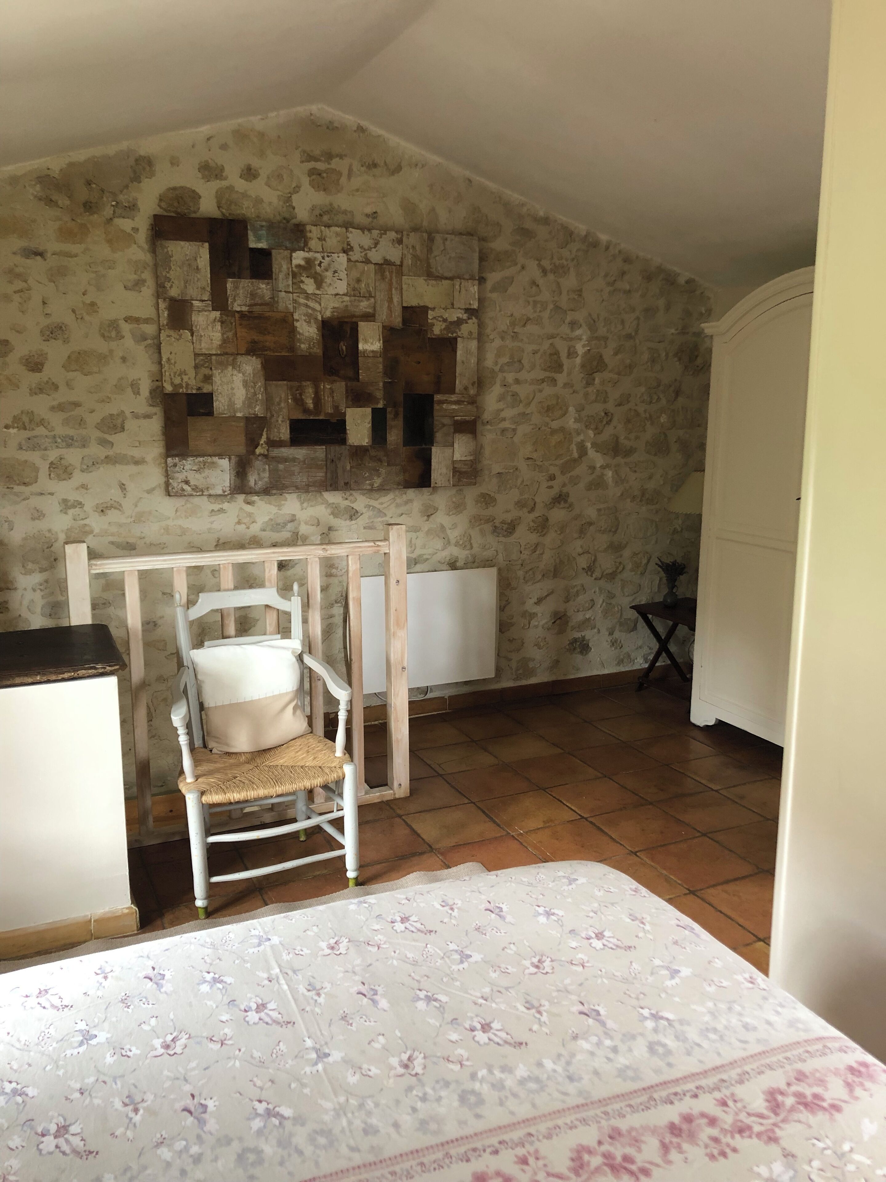 Une pièce avec un mur en pierre rustique, une chaise en bois avec un coussin, un radiateur et un lit recouvert d’une couvre-lit à motifs floraux.