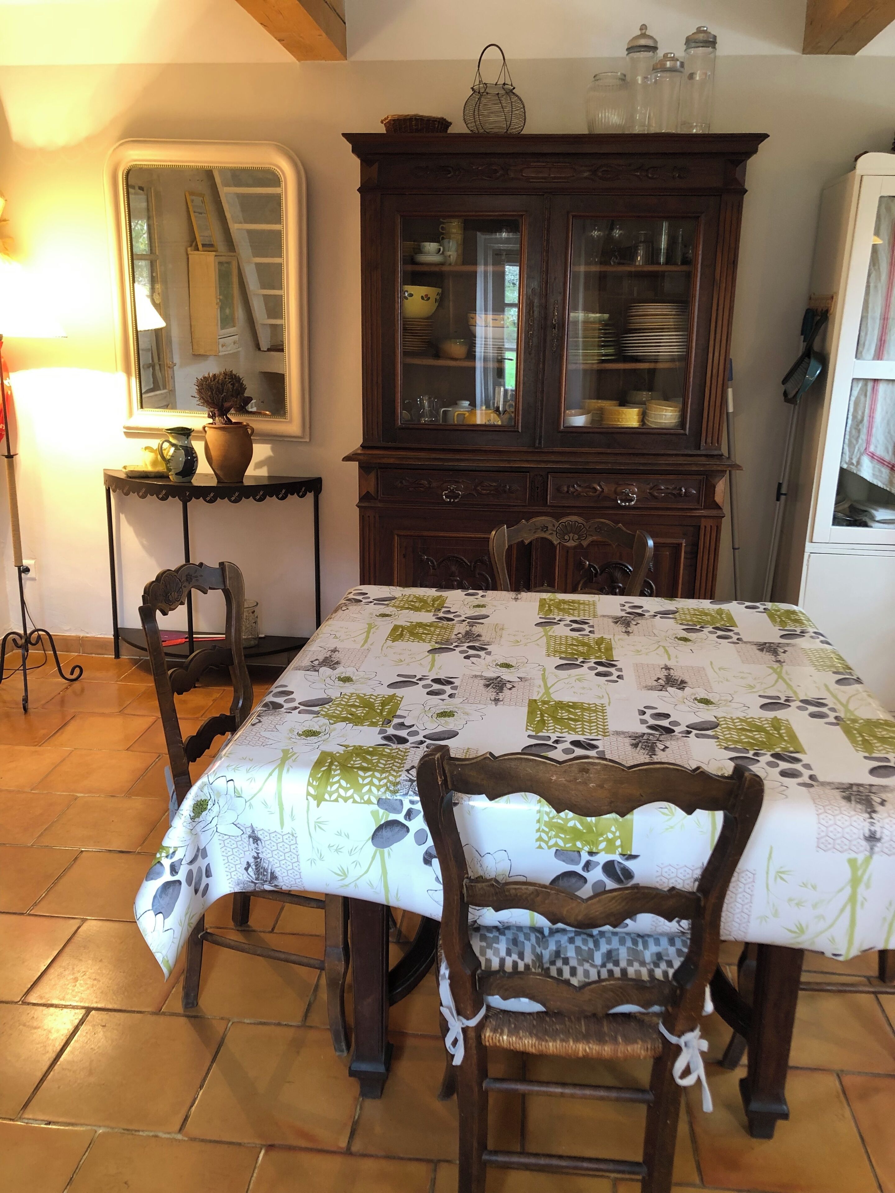 A dining area with a table covered in a floral tablecloth, surrounded by wooden chairs. A dark wooden cabinet with glass doors is in the background, and a mirror hangs on the wall.
