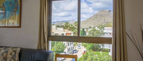 A room with a large window offering a view of a mountainous landscape and a town. There is a wooden table with a plate and a chair.