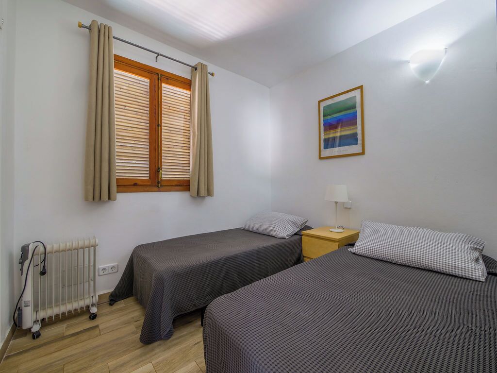 A room with two single beds, a radiator, a window with curtains, a nightstand with a lamp, and a framed picture on the wall.
