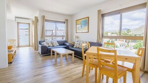 Un salon spacieux comprenant un canapé, une table basse et une table à manger. De grandes fenêtres offrent une vue sur l’extérieur.