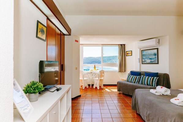 A hotel room with a bed, a sofa, a small table with chairs, a TV, and a window with a view of the sea.