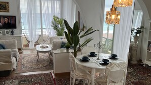 Een ruime woonkamer met een eettafel gedekt voor vier personen, omringd door witte stoelen. Een grote plant staat in het midden, en de kamer heeft een hoog rondboogvenster met lichte gordijnen.
