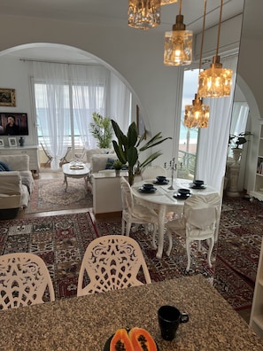 A spacious living area with a dining table set for four, surrounded by white chairs. A large plant is in the center, and the room features a high arched window with sheer curtains.