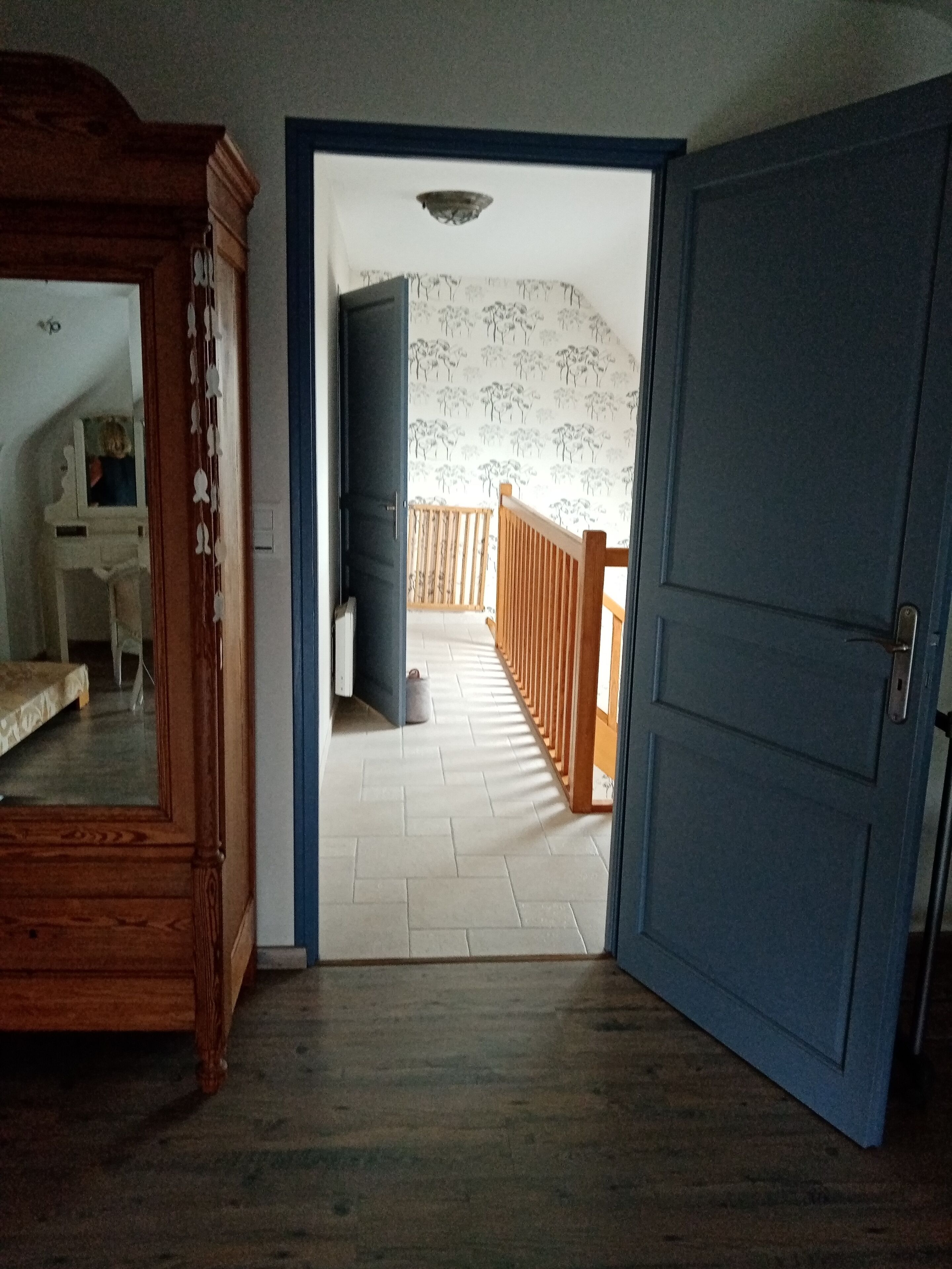 A room with a wooden wardrobe, a bed, and a door leading to a balcony with a view of trees.