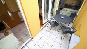 Pequena varanda com mesa redonda e quatro cadeiras, porta de correr de vidro e vista para outra varanda com sofá e mesa.