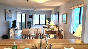 A dining area with a wooden table, chairs, and a chandelier. There is a television and a plant in the background. - Bright, corner unit, with open floor plan and views from nearly every room!  (Bradenton Beach, Anna Maria Island)