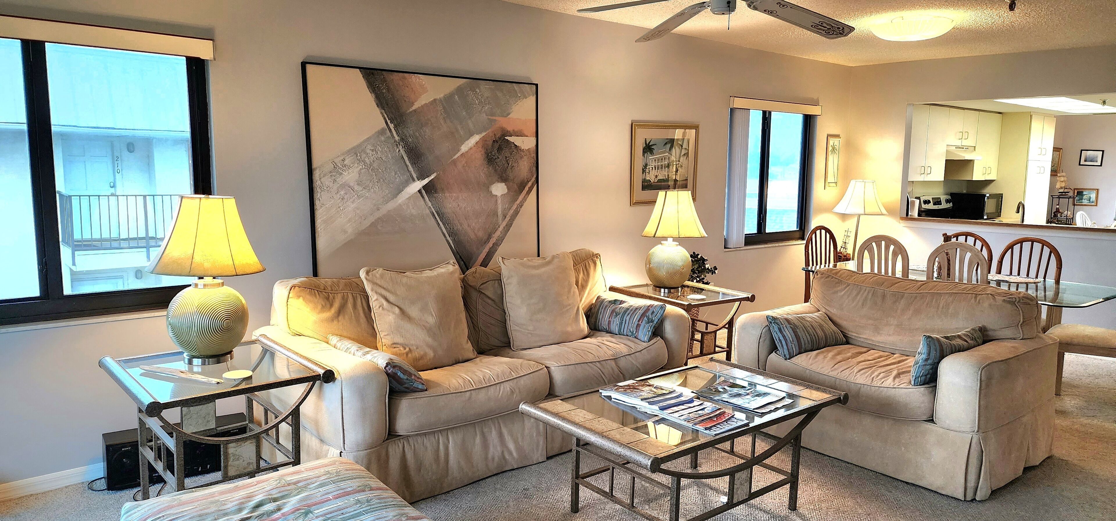 Ein Wohnzimmer mit zwei Sofas, einem Couchtisch und einem Essbereich mit Stühlen. An der Wand hängt ein großes abstraktes Gemälde, und durch eine offene Tür ist eine Küche zu sehen.